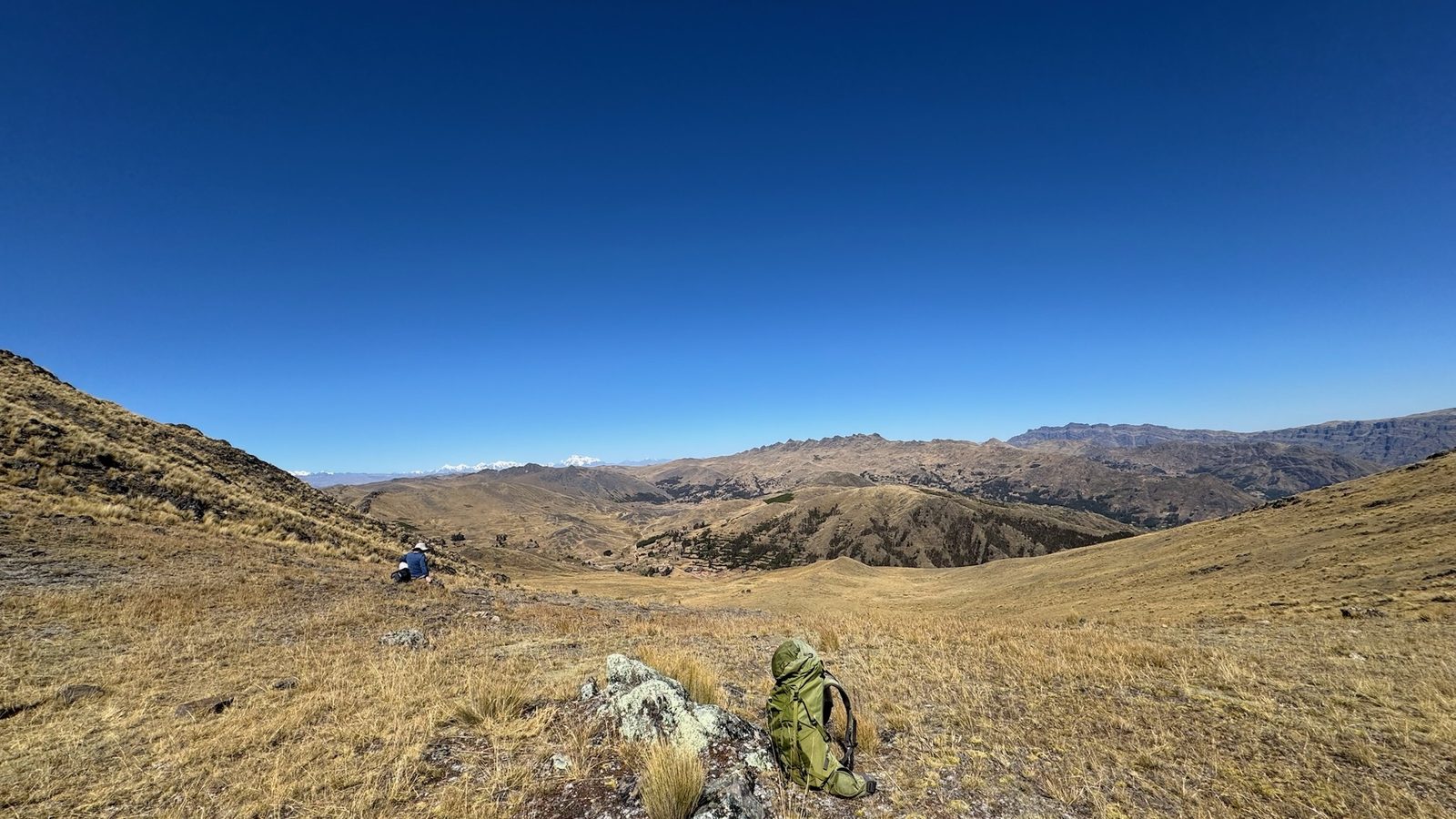 A wide valley of golden grassland falling away between bare hills to snow peaks on the far horizon, a rucksack resting on a lichened rock in the foreground