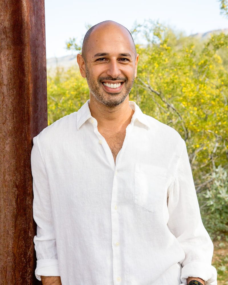 Hamid Jabbar standing outdoors in an open white linen shirt, smiling, beside a weathered steel post with flowering desert trees behind him