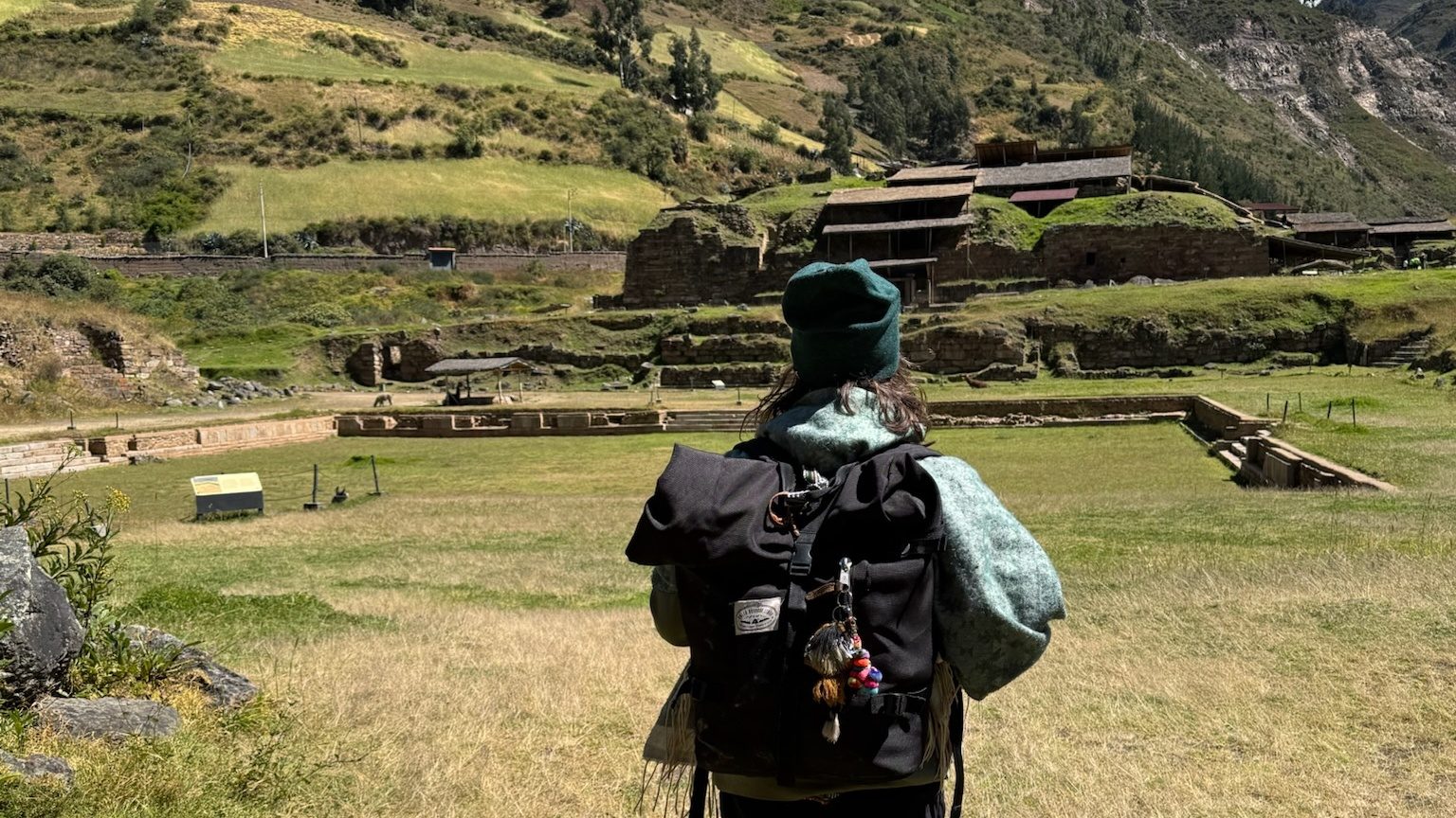 Someone seen from behind looking across grass to the stepped stone terraces of a ruined temple complex, green mountains rising behind