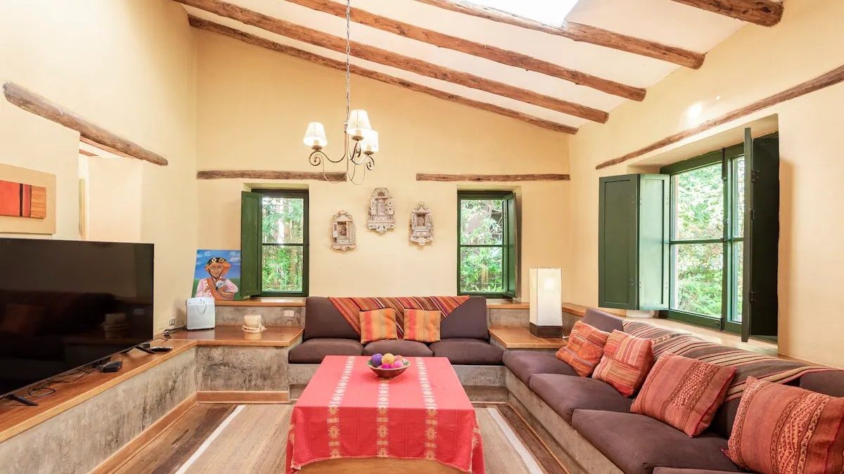 A sitting room under a beamed ceiling with a long low sofa, red and orange cushions, a red woven cloth on the table and green framed windows onto the garden
