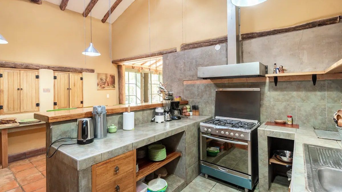 A kitchen with a concrete counter, a gas range under a steel hood, open wooden shelves and a window onto the garden