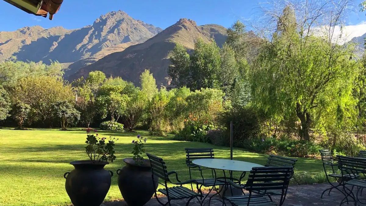 A stone terrace set with metal garden chairs and two planted urns, looking across a lawn and trees to a bare mountain