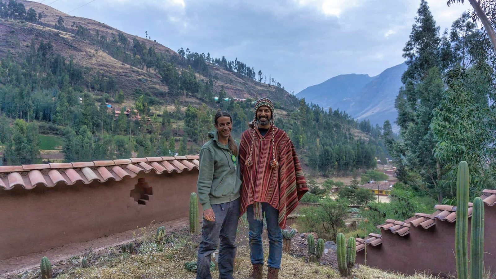 Two people standing together outside an adobe house, with cacti and mountains behind them